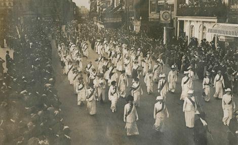 A black and white photo of a number of women marching in the street, wearing matching white outfits and dark sashes while crowds look on